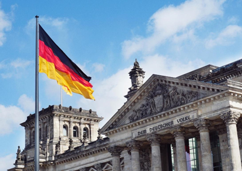 Die deutsche Flagge und im Hintergrund das Reichstagsgebäude in Berlin.  Quelle: https://unsplash.com/photos/HF-lFqdOMF8
